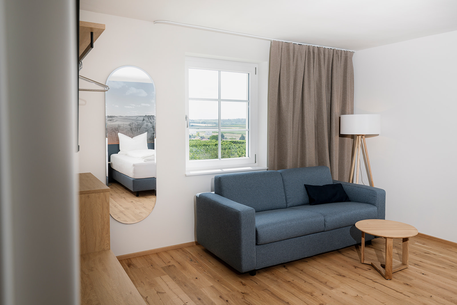 Heller Wohnbereich mit blauem Schlafsofa, Spiegel und Blick auf die Weinviertler Landschaft im Zollquartier